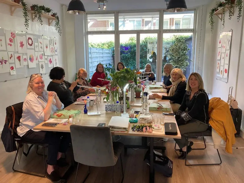 A group of women gathered around a long table in a bright, modern room with large windows, decorated with greenery and framed artwork. The table is filled with notebooks, pens, and other office items, suggesting a workshop, meeting, or class setting.
