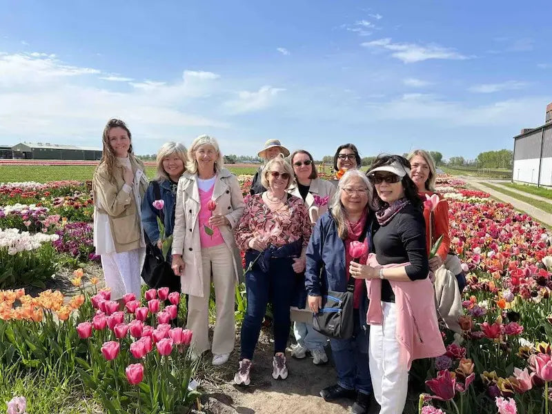A group of nine women posing together in a vibrant tulip field under a blue sky. They are dressed in casual spring attire, with pink tulips lining the path on both sides.