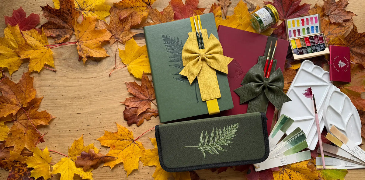 Green fern watercolor sketchbook with yellow bow brush holder, red Bow Sketchbook, Forest Brush Case and ceramic palette on autumn leaves